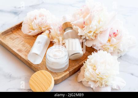 Creme, Serum, Essenz, Öl in Milchglasverpackung mit rosa Pfingstrosen-Blüten. Kosmetische Hautpflege Produkt aus Holz Tablett. Ausgewählter Fokus. Stockfoto