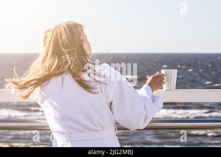 Eine Frau in einem weißen Gewand mit einem weißen Becher in den Händen auf der Terrasse blickt auf das Meer, eine romantische Stimmung. Stockfoto