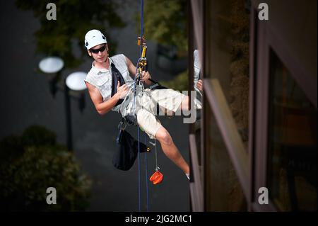 Industriebergsteiger Arbeiter in Schutzhelm zeigt Genehmigung Geste Daumen nach oben, während an Kletterseil vor dem Gebäude hängen. Mann in Sonnenbrillen, der Sicherheitsausrüstung im Freien verwendet. Stockfoto