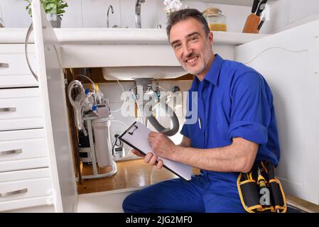 Sanitär-Techniker prüft Wasserinstallation unter dem Waschbecken einer Wohnküche mit Notepad. Stockfoto