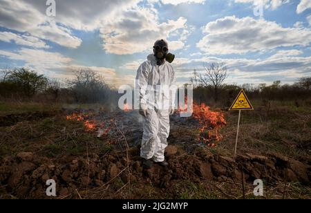 Feuerwehrmann-Ökologe, der mit einem Waldbrand auf dem Feld arbeitet. Mann in Gasmaske neben brennendem Gras mit Rauch und gelbem Dreieck mit Totenkopf und Kreuzknochen Warnschild. Konzept für Naturkatastrophen. Stockfoto