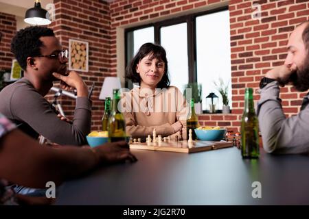 Fröhliche Frau, die mit Freunden zu Hause im Wohnzimmer sitzt, während sie Schach spielt. Fröhliche und vielfältige Menschen genießen gemeinsam strategische Brettspiele, während sie sich entspannen und Snacks und Getränke konsumieren. Stockfoto