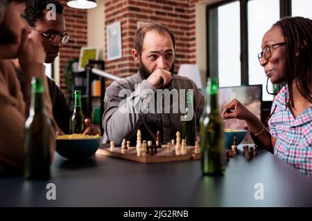 Nachdenklicher Mann, der im Wohnzimmer mit multiethnischen Freunden am Tisch sitzt, während er Schach spielt. Angespannte Person genießt Strategie-Brettspiele, während sie über Schachbrett nächsten Schritt denken. Stockfoto