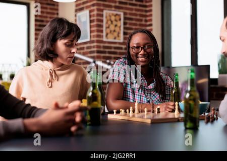 Multiethnische Gruppe von Freunden, die zu Hause zusammen sitzen, während sie Schach spielen. Glückliche, kluge Menschen im Wohnzimmer genießen strategische Brettspiele und konsumieren Snacks und Getränke. Stockfoto