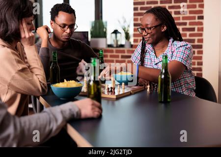 Schlaue Leute, die im Wohnzimmer am Tisch sitzen und gemeinsam Schach spielen. Fröhliche, fröhliche, multiethnische Freunde sitzen zu Hause zusammen und genießen Strategiespiele, Snacks und Getränke. Stockfoto