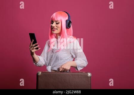 Weibliches Modell mit Smartphone und Koffergepäck vor der Studiokamera. Reisen Sie auf Städtereise und bereiten Sie sich auf eine Abenteuerreise und eine Reise mit dem Aktentaschenwagen vor. Stockfoto