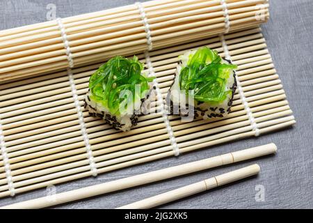 Seetang-Sushi auf einer Bambusmatte. Essstäbchen auf dem Tisch. Draufsicht. Grauer Hintergrund. Stockfoto