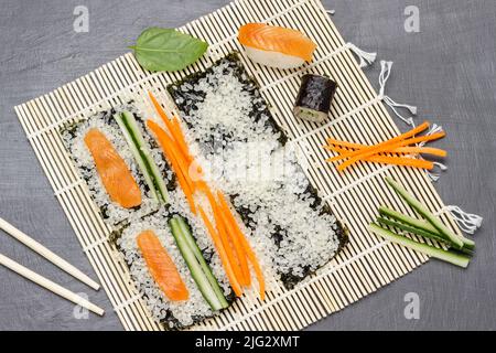 Prozess der Herstellung von Sushi. Reis, gehackte Karotten und Gurken sowie Lachsscheiben auf einer Bambusmatte. Flach liegend. Grauer Hintergrund. Stockfoto