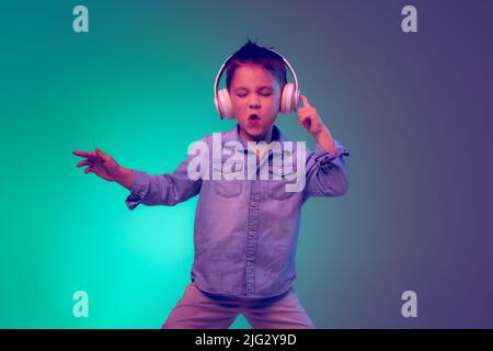 Porträt eines gefühlvollen kleinen Jungen, Kind, das Musik in Kopfhörern hört, isoliert auf grün-violettem Hintergrund in Neonlicht posiert Stockfoto