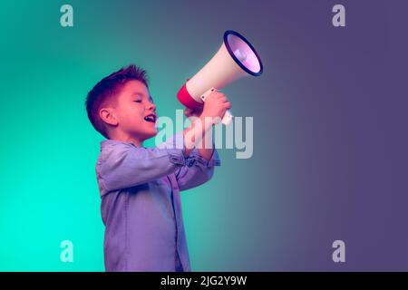 Porträt eines fröhlichen kleinen Jungen, Kind schreiend in Megaphon, posiert isoliert auf grünem lila Hintergrund in Neonlicht Stockfoto