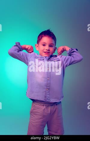 Porträt des kleinen Jungen, Kind in Jeans T-Shirt posiert isoliert auf grün lila Hintergrund in Neonlicht. Verspieltes Kind Stockfoto