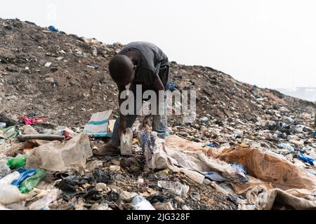 Ein nicht erkennbarer Jugendlicher in einem getragenen schwarzen T-Shirt und einer zerfetzten Hose auf der Suche nach Gegenständen im Müll, der auf einer großen Deponie in Westafrika verschüttet wurde Stockfoto