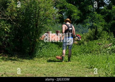 Bauer mit Strohhut mäht hohes Gras in einem Hinterhof hinter dem Haus im Dorf an heißen Sommertagen. Prozess des Rasentrimmens. Gartenkonzept Stockfoto