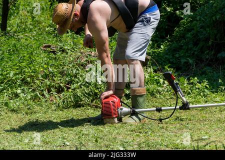 An einem sonnigen Sommertag startete der Bauer in einem Hinterhof einen Handrasen-Benzinmäher. Gartengeräte- und Ausrüstungskonzept. Nahaufnahme, selektiver Fokus Stockfoto