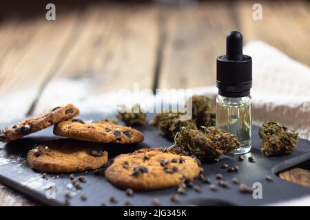 Hausgemachte Chocolate Chip Cookies mit cbd-Öl, umgeben von trockenen Cannabisknospen, Marihuanasamen und einer Glasflasche mit einer Pipette CBD-Öl. Stockfoto