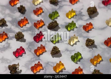 Mehrere trockene Knospen von medizinischem Marihuana liegen in der Reihenfolge unter Gummibären verschiedener Farben mit Zusatz von cbd-Öl auf einem Marmortisch, Stockfoto