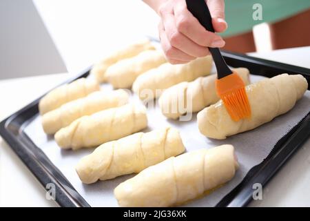 Der Koch bürstet rohe Croissants mit der Bürste im Eigelb-Kochvorgang Stockfoto