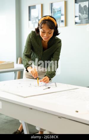 Lächelnde asiatische junge Architektin, die Blaupause am Tisch im Kreativbüro zeichnet Stockfoto