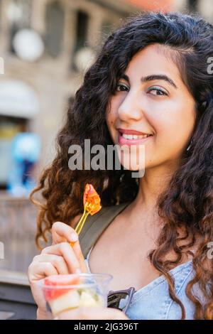 türkische junge Frau, die die Kamera anschaut, während sie einen Plastikbecher mit Fruchtstücken in der Hand hält Stockfoto