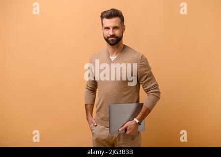 Waist-up-Porträt des lächelnden Mannes, der Laptop in den Händen hält und lächelt, ein gutaussehender freiberuflicher Mann, der isoliert auf beigem Hintergrund steht, Kopierraum Stockfoto