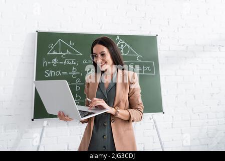 Lächelnder Lehrer, der in der Schule einen Laptop in der Nähe von mathematischen Formeln auf der Tafel verwendet Stockfoto