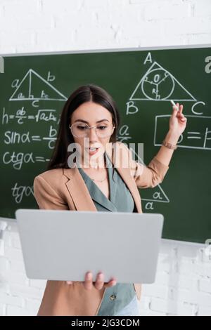 Lehrer mit Videoanruf auf Laptop in der Nähe der Tafel mit mathematischen Formeln Stockfoto