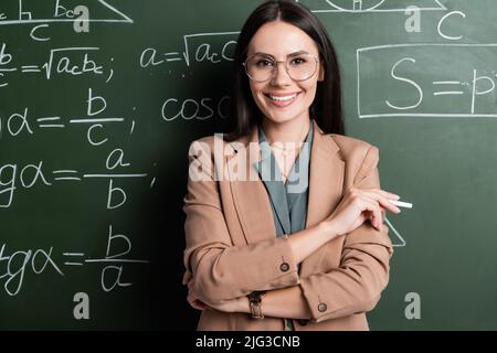 Positiver Lehrer, der Kreide in der Nähe von mathematischen Formeln auf dem Schwarzen Brett hält Stockfoto