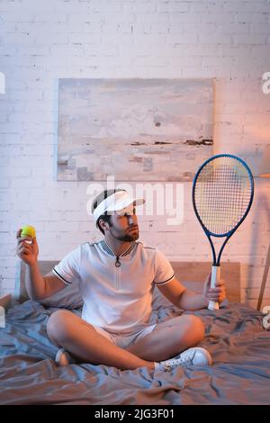 Verwirrte Schlafwandlerin in Sportkleidung, die Tennisball und Rakete auf dem Bett hielt Stockfoto