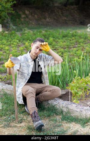 Vertikale Ansicht des jungen Gärtners müde sitzen mit einer Hacke nach der Arbeit auf dem Garten Gemüseanbau Stockfoto