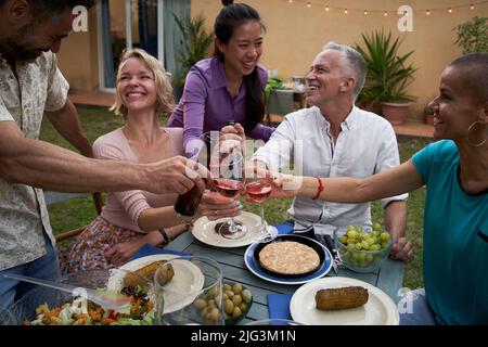 Eine Gruppe von Freunden toasten mit Wein beim Grillen im Hinterhof. Glückliche Menschen mittleren Alters, die sich bei einem Picknick im Garten amüsieren. Stockfoto