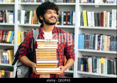 Intelligenter positiver indischer oder arabischer Tausendjähriger, Student, in lässiger Kleidung, steht in der Bibliothek, hält viele Bücher in seinen Händen, bereitet sich auf die Prüfung vor, schaut weg, lächelt glücklich Stockfoto