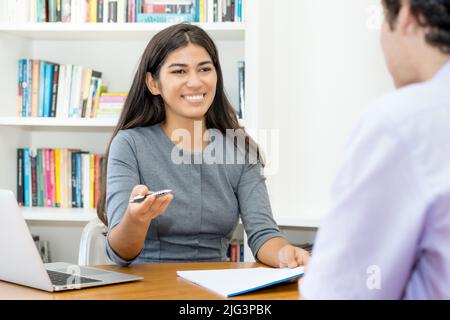 Lachender südamerikanischer Business-Trainee im Gespräch mit einem Kollegen im Büro Stockfoto