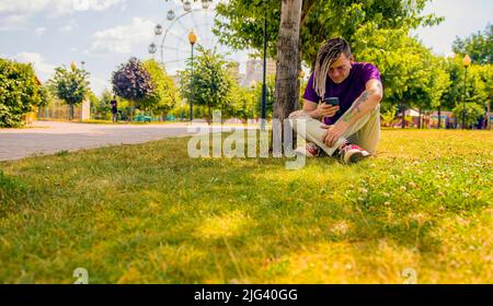 Ein junger Mann mit Dreadlocks ruht im Schatten eines Baumes und nutzt ein Smartphone im Park. Ein gutaussehender Kerl mit Sonnenbrille sitzt auf dem Gras Stockfoto