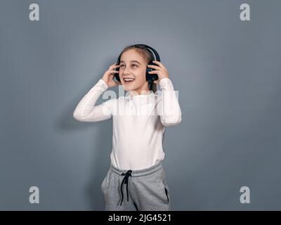 Energiegeladenes kleines Mädchen in Kopfhörern. Weißes T-Shirt, graue Jogginghose. Hält Kopfhörer, hört Musik Stockfoto