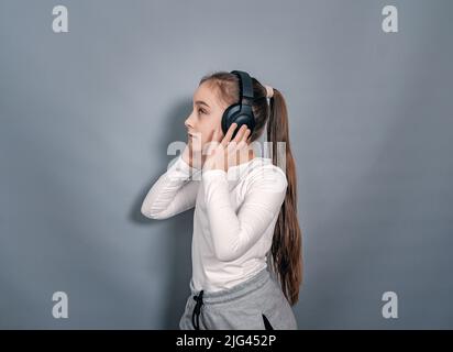 Seitenansicht kleines Mädchen in Kopfhörern. Weißes T-Shirt, graue Jogginghose. Hält Kopfhörer, hört Musik Stockfoto