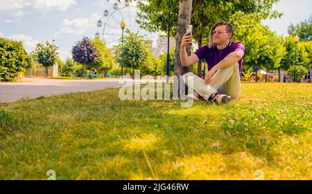 Ein junger Mann mit Dreadlocks ruht im Schatten eines Baumes und nutzt ein Smartphone, um ein Selfie im Park zu machen. Ein gutaussehender Kerl in Sonnenbrille sitzt Stockfoto