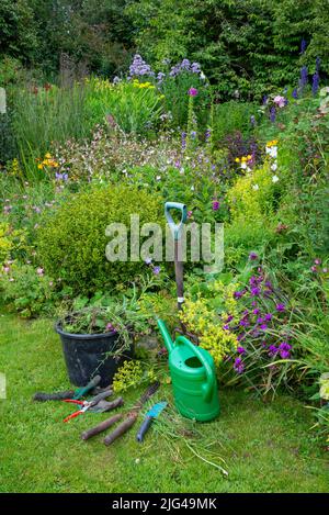 Englischer Landgarten Anfang Juli. Eine Auswahl an gut verwendeten Werkzeugen neben den Grenzen voller verschiedener Pflanzen. Stockfoto