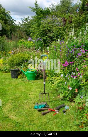 Englischer Landgarten Anfang Juli. Eine Auswahl an gut verwendeten Werkzeugen neben den Grenzen voller verschiedener Pflanzen. Stockfoto