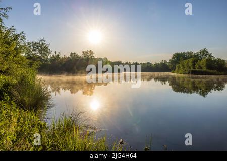 Morgennebel kriecht mit Sonnenreflexion über die Wasseroberfläche des Sees Stockfoto