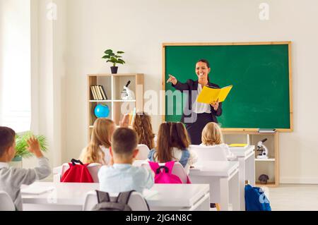 Freundliche Lehrerin im Klassenzimmer mit Buch in den Händen unterrichtet Grundschüler. Stockfoto