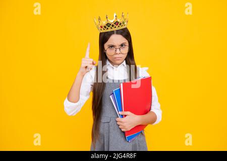 Schulmädchen in Schuluniform und Krone feiern den Sieg auf gelbem Hintergrund. Schulkind hält Bücher. Bildungsabschluss, Sieg und Erfolg Stockfoto