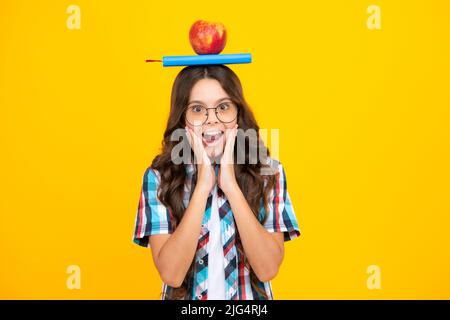 Zurück zur Schule. Teenager Schülerin mit Apfel auf dem Kopf, bereit zu lernen. Schulkinder auf isoliertem gelben Hintergrund. Schockiert überrascht Teenager Mädchen Stockfoto