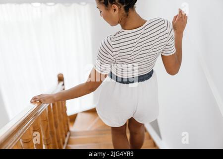 Junge schwarze Frau in Hemden, die zu Hause die Holztreppe hinuntergeht Stockfoto