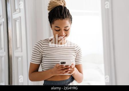 Junge schwarze Frau mit Zöpfen lächelnd und mit Handy zu Hause Stockfoto