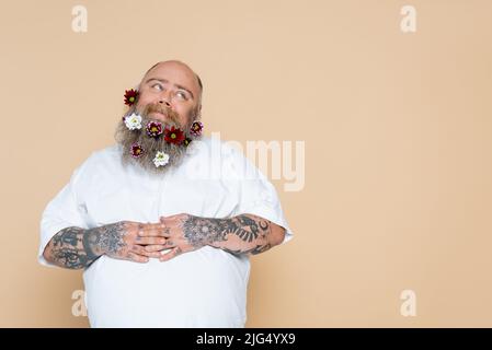 Zufrieden übergewichtiger Mann mit Blumen im Bart und Hände auf Bauch isoliert auf beige Stockfoto