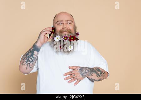 Fröhlicher Mann mit Blumen im Bart, der auf einem Mobiltelefon auf Beige isoliert spricht Stockfoto