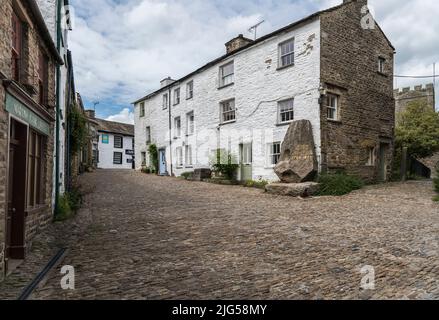 Gebäude und leere gepflasterte Straße im Zentrum von Yorkshire Dales Dorf mit Denkmal für Adam Sedgwick und einem castellierten Kirchturm im Hintergrund. Stockfoto