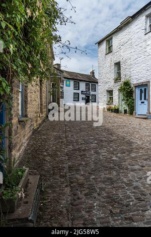 Leere gepflasterte Straße im Dorf Yorkshire Dales mit Sträuchern im Vordergrund und Sun Inn im Hintergrund unter blauem Himmel mit weißen Wolken. Stockfoto