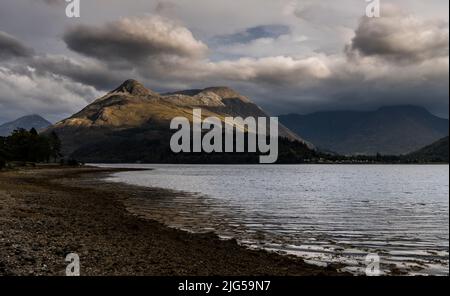 Abendblick über das helle Wasser von Loch Leven zum sonnenbeschienenen Pap of Glencoe mit den Bergen am westlichen Ende von Glencoe im Schatten unter niedriger Wolke Stockfoto