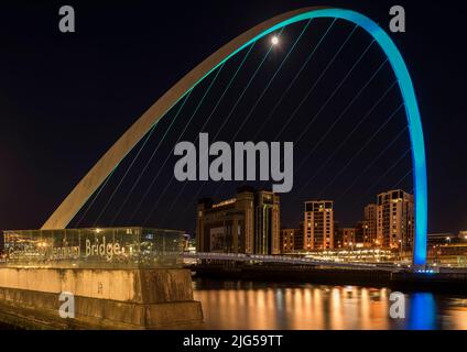 Die Gateshead Millennium Bridge wurde nachts mit einem Flutlicht beleuchtet, das Baltic Arts Center und andere Gebäude mit Flutlicht im Hintergrund und Vollmond, der durch den Bogen leuchtet. Stockfoto